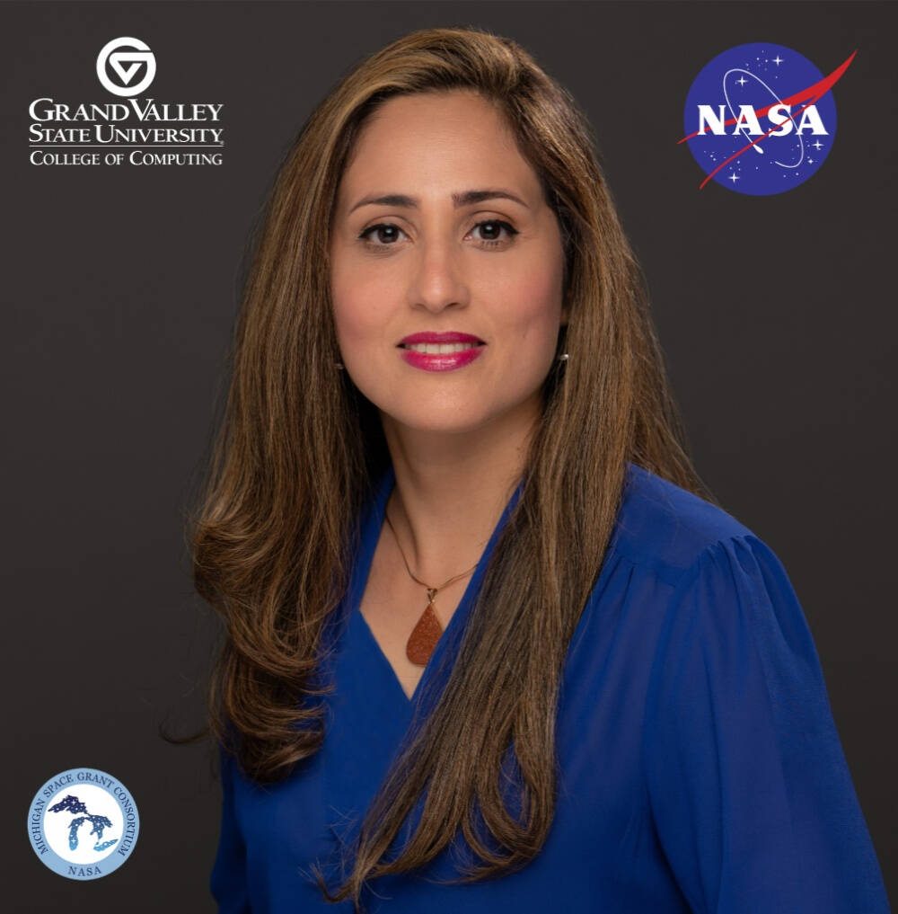 Dr. Sara Sutton wearing a blue chiffon top is staring at a  camera in front of a gray GVSU College of Computing branded cover drop. At the top right, there's a NASA logo. She has long brown hair.